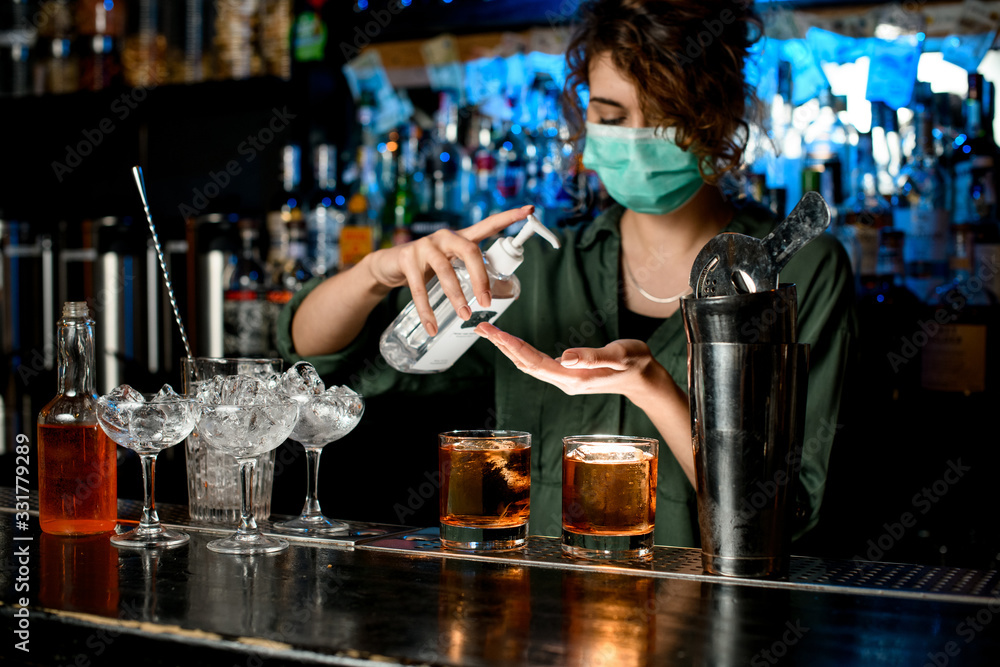 Woman in medical mask treats her hands with disinfector behind bar counter.