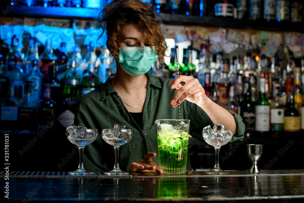 woman bartender in medical mask mixes green drink in glass using spoon.