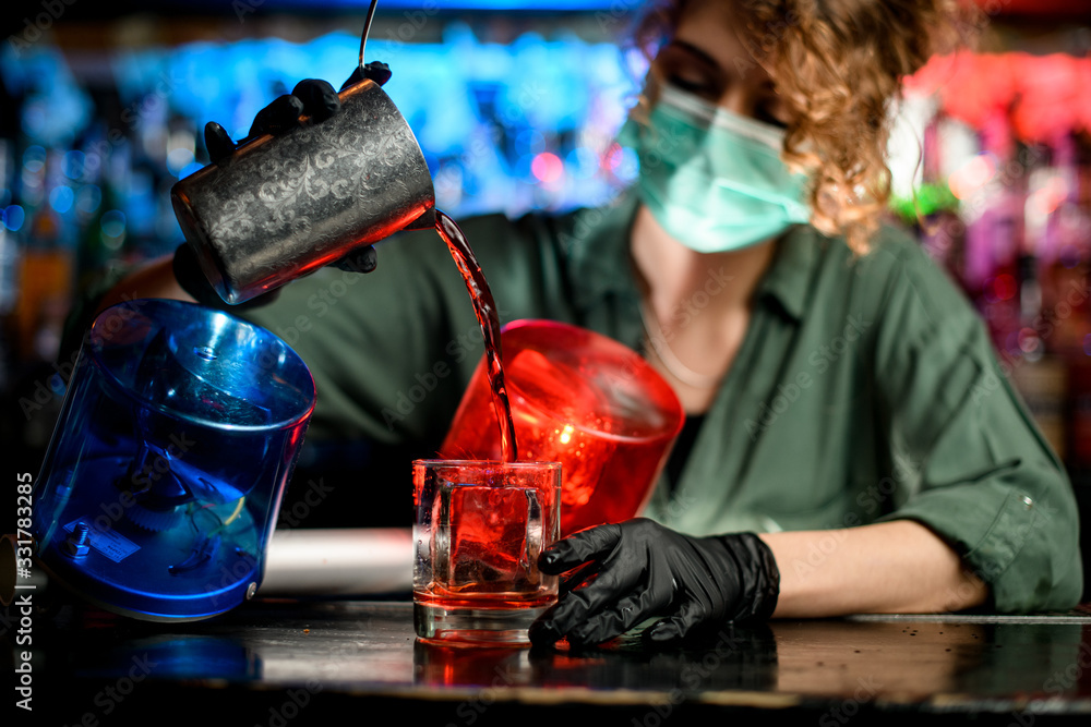 Young bartender girl in medical mask and black gloves professionally pour drink from steel glass into glasses with ice. - obrazy, fototapety, plakaty 