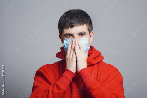 Man in medical mask coughing and covers his mouthon gray studio backround, concept of prevention viral deseases, person in red sweatshirt with dangerous symptoms