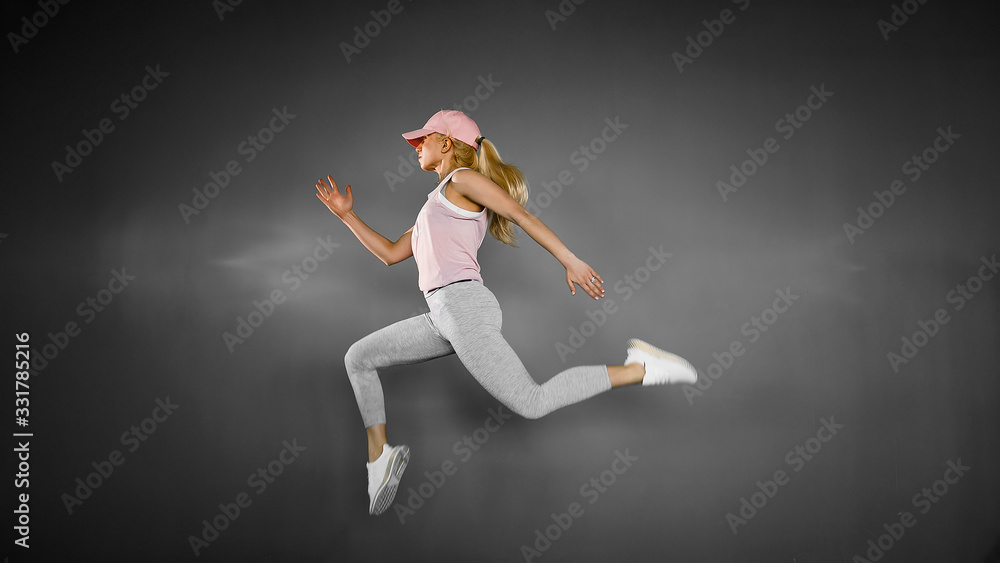 Young blonde woman with fit body jumping and running against grey background. Female model in sportswear exercising.