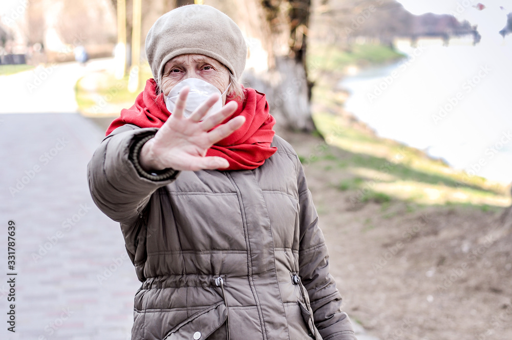 Old woman in a protective mask on her face and outstretched arm shows a stop sign on the deserted embankment of the city. COVID-2019 older people. Coronavirus Affects Senior Health - Keeping distance. Old woman in a protective mask on her face and outstretched arm shows a stop sign on the deserted embankment of the city. COVID-2019 older people. Coronavirus Affects Senior Health - Keeping distance.