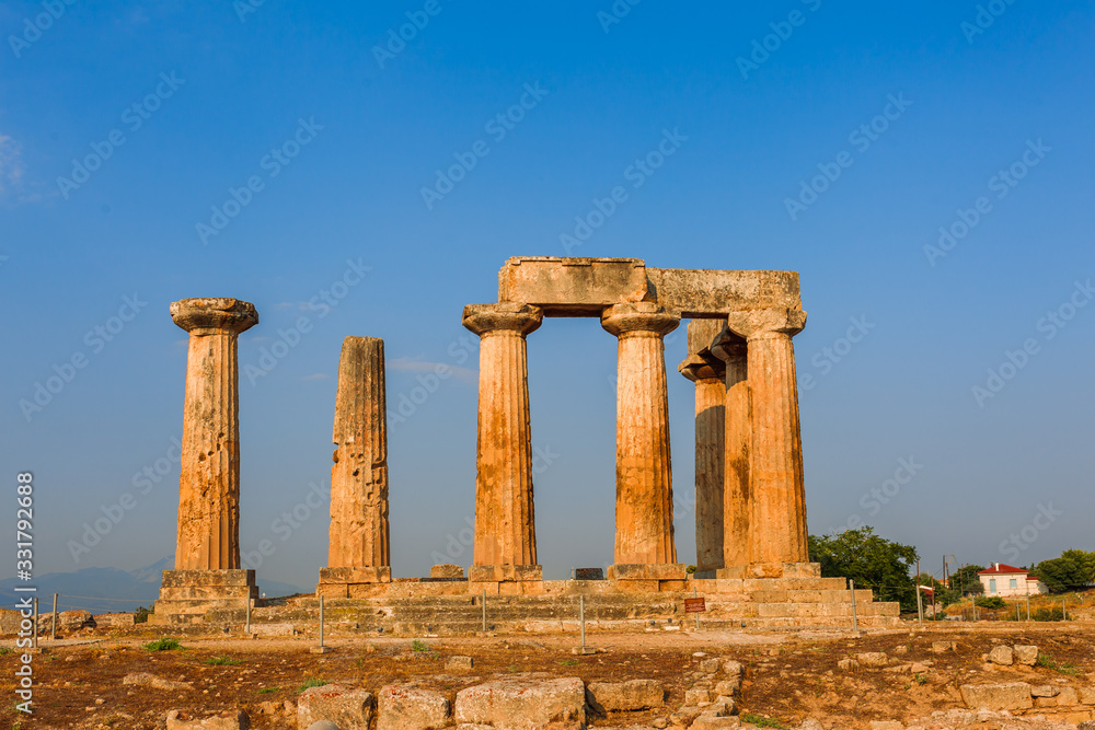 Ruins of Temple of Apollo in Corinth Greece standing up on a hill with remenants of rock walls scattered about under a bright sun with mountians and a blue sky behind