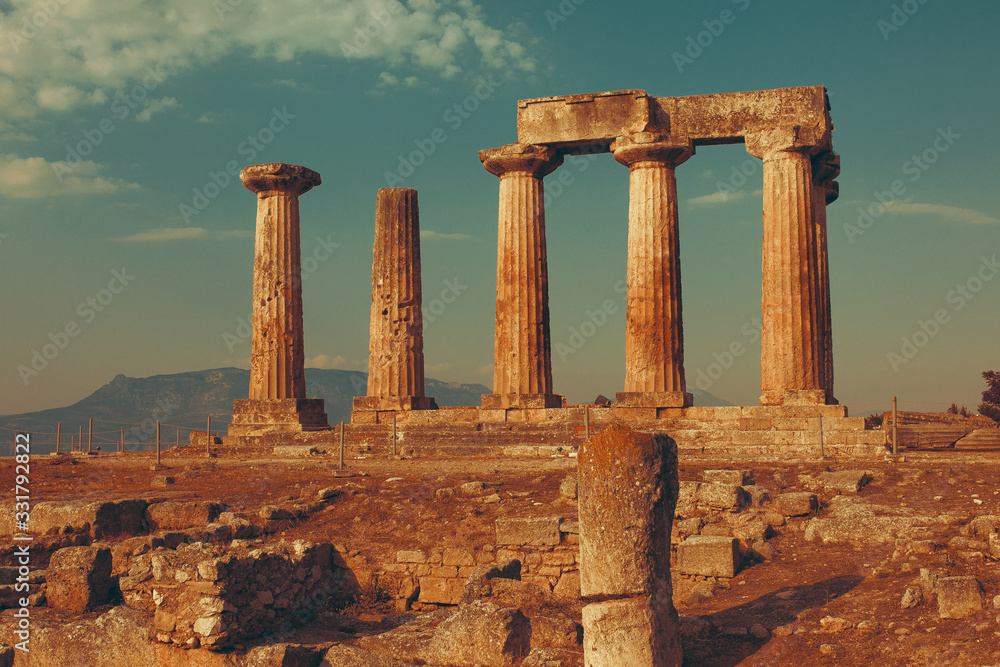 Fototapeta premium Ruins of Temple of Apollo in Corinth Greece standing up on a hill with remenants of rock walls scattered about under a bright sun with mountians and a blue sky behind