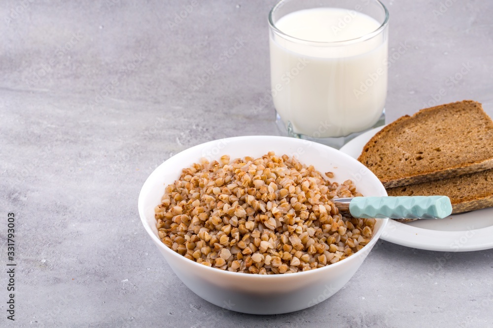Cooked buckwheat in white bowl with spoon, glass of milk, piece of bread on grey background. Close up, space for text, horizontal format. Cooked buckwheat in white bowl with spoon, glass of milk, piece of bread on grey background. Close up, space for text, horizontal format.