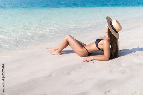 Beautiful woman with perfect body lying down on the beach, wearing stylish hat, tanning on a beach resort, enjoying summer vacation
