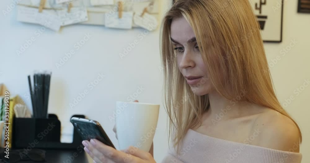 Portrait young blonde woman leafs through social media feed on smartphone over drink cup of coffee, and smiles, sits in coffee shop behind counter by window, slide. Chatting in Internet. Lifestyle Portrait young blonde woman leafs through social media feed on smartphone over drink cup of coffee, and smiles, sits in coffee shop behind counter by window, slide. Chatting in Internet. Lifestyle