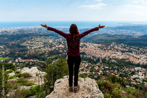 Full body shot of young Caucasian woman standing on top of cliff enjoying the view of Vence and other towns, vast French Alps mountains and the Mediterranean Sea landscape with her hands in the air