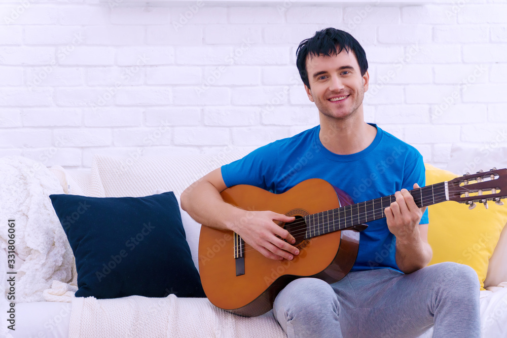 Handsome young guy playing acoustic guitar while sitting on sofa at white living room. Attractive man holding guitar in his hands felling relaxation while looking at camera at home. Lifestyle concept.
