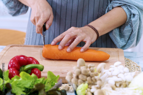 Close-up hand of woman wear apron sliced carrots with steel knife on wooden chopping block with mushrooms and vegetarian background while cooking healthy food in the kitchen. Vegetarian food concept.