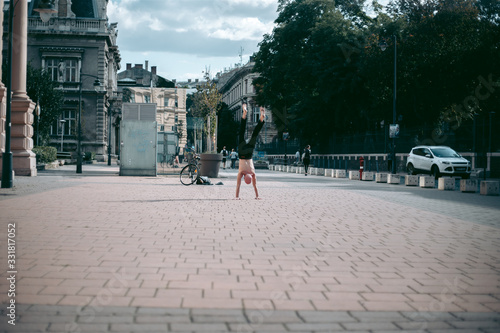 An old bald man with a naked torso handstands in the city center