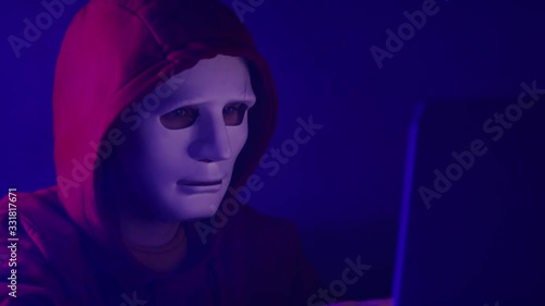 Young anonymous hacker man in red hoodie and mask working on the laptop in the room filled with smoke. Close up slow motion shot photo