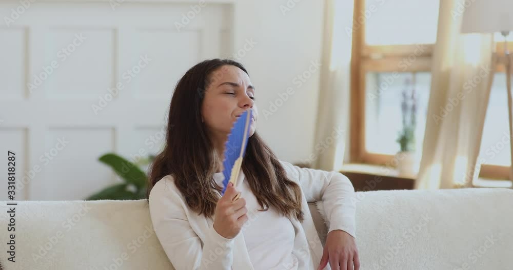 Exhausted frustrated young lady feeling uncomfortable hot waving fan sitting on sofa in sultry apartment. Annoyed overheated woman suffering from summer heat or broken air conditioner indoors. Exhausted frustrated young lady feeling uncomfortable hot waving fan sitting on sofa in sultry apartment. Annoyed overheated woman suffering from summer heat or broken air conditioner indoors.