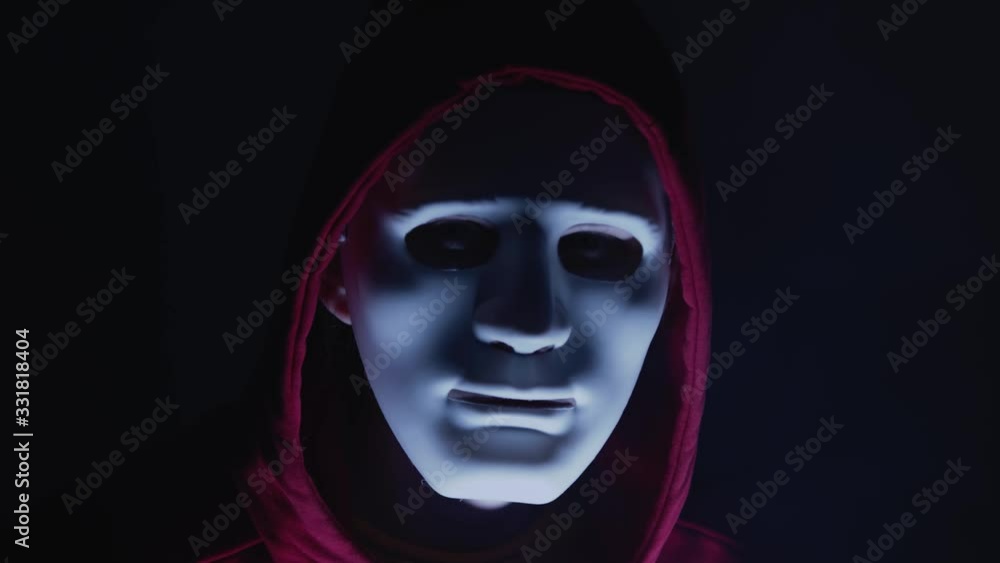Scary hacker with mask looking to the camera. Lights flashing around him. Anonymous man coding in the dark room Scary hacker with mask looking to the camera. Lights flashing around him. Anonymous man coding in the dark room