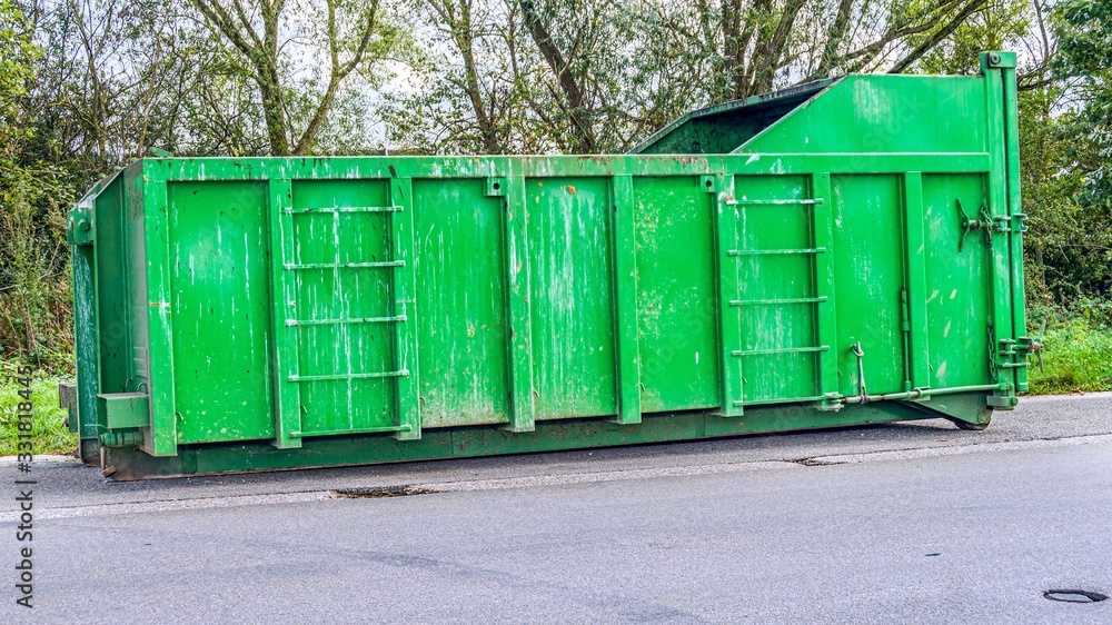 Disposal and recycling dumpster . A large, metal, green garbage container and municipal waste, standing on a dirt road near the fence and trees. 