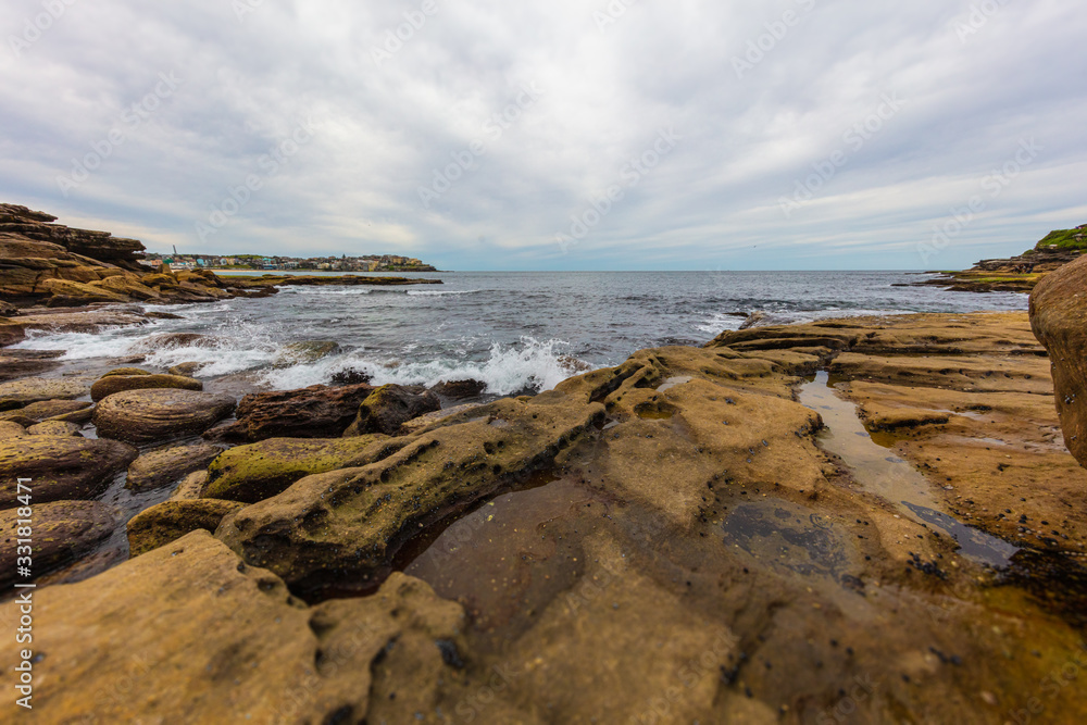 The rocky shoreline around Bondi beach, Sydney, Australia. Waves splashing at the rocks in the water. White clouds on blue sky. The cliffs beside the sandy beach. A paradise for surfers 