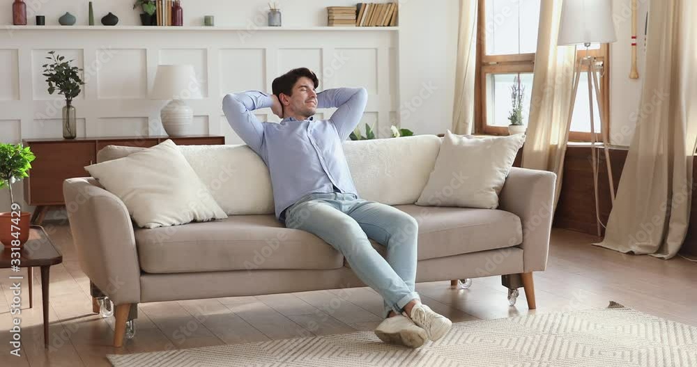 Smiling young man relaxing in modern living room sitting on comfortable sofa in cozy apartment. Happy lazy guy holding hands behind head enjoying lounge daydreaming on weekend stress free day at home. Smiling young man relaxing in modern living room sitting on comfortable sofa in cozy apartment. Happy lazy guy holding hands behind head enjoying lounge daydreaming on weekend stress free day at home.
