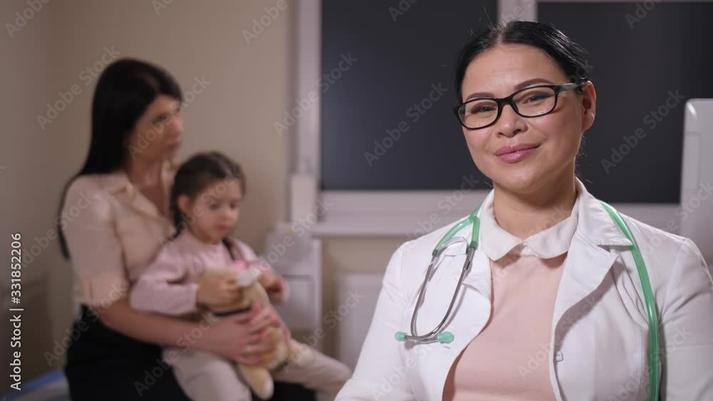 Close-up portrait of friendly asian pediatrician in glasses posing in own office, little girl patient sitting on mom's knees blurred on background. Confident mature family doctor smiling at camera Close-up portrait of friendly asian pediatrician in glasses posing in own office, little girl patient sitting on mom's knees blurred on background. Confident mature family doctor smiling at camera