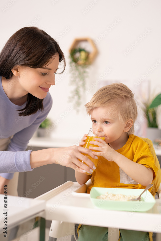 Smiling young mother standing at highchair and giving fresh juice to little son in kitchen