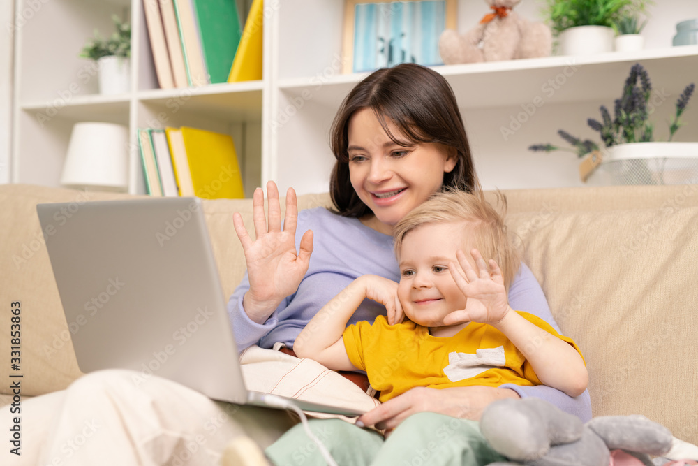 Cheerful young mother and son sitting on sofa and waving hands while talking to father via video chat Cheerful young mother and son sitting on sofa and waving hands while talking to father via video chat