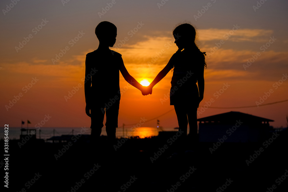 Children hold hands in the background of the sunset. Silhouettes of a young boy and girl.