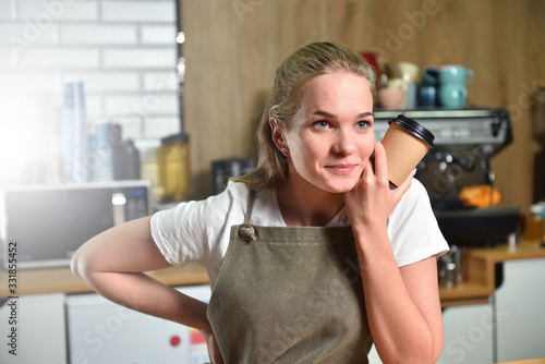 A young barista girl holds a paper cup with coffee in her hands and smiles thoughtfully. Small business concept