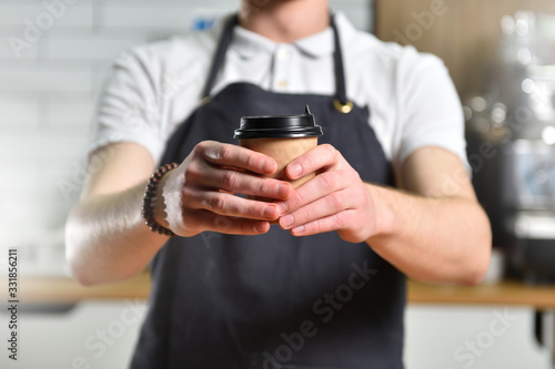 Young guy bartender barista holds out his hand with a paper cup of takeaway coffee. Small Business Concept