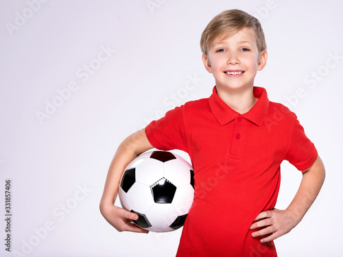 Photo of a smiling boy in sportswear holding soccer ball, posing at studio. Happy 8 years old kid in a red t-shirt with a soccer ball in hand. White child with a smile holds a soccer ball.