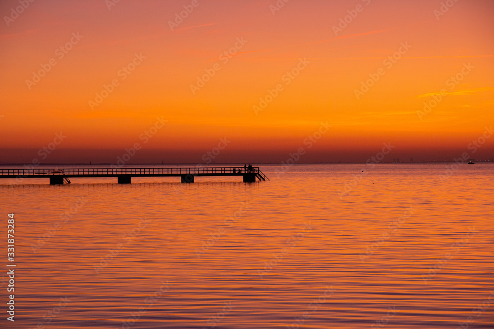 People have walked out on the pier in Malmö, Sweden, while the sun is setting and making the sea and sky all orange and pink. People have walked out on the pier in Malmö, Sweden, while the sun is setting and making the sea and sky all orange and pink.