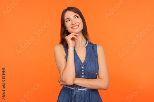 Smiling happy brunette woman in denim dress looking up with dreamy peaceful expression and imagining, thinking of pleasant memories, making wish. indoor studio shot isolated on orange background
