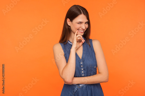 Fotografija Beautiful and sly brunette woman in denim dress pondering mysterious tricky plan, smiling with cunning playful expression, scheming evil plan, devious idea