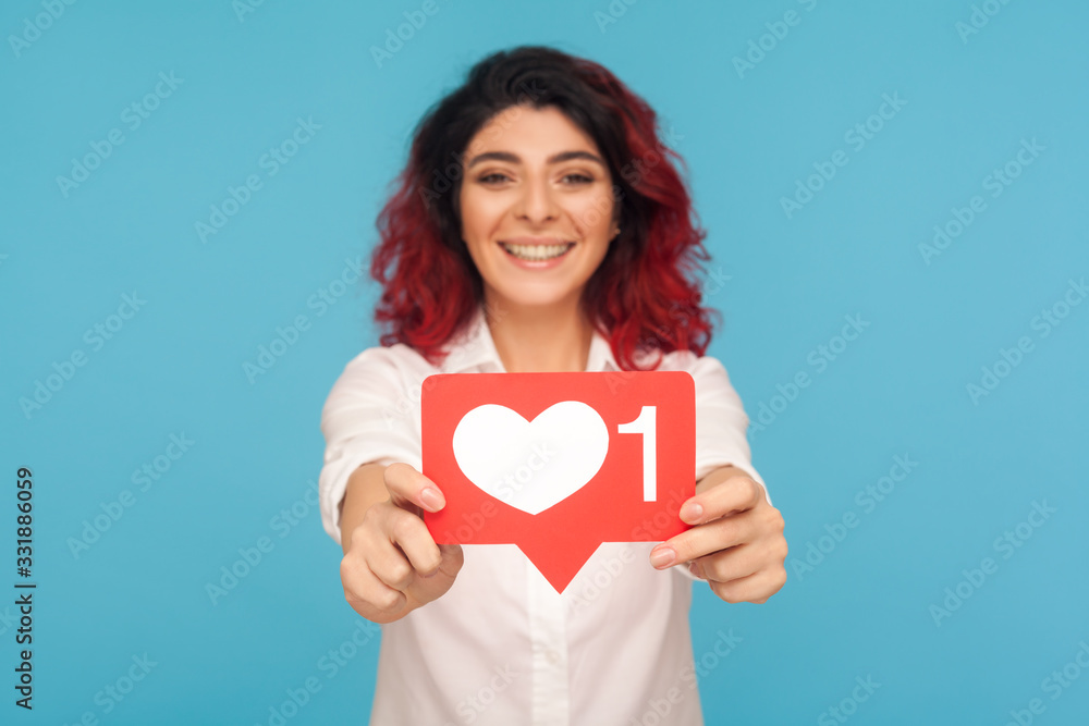 Portrait of beautiful woman with fancy red hair showing social media heart Like icon, symbol of popularity in internet, recommending to push button. indoor studio shot isolated on blue background Portrait of beautiful woman with fancy red hair showing social media heart Like icon, symbol of popularity in internet, recommending to push button. indoor studio shot isolated on blue background