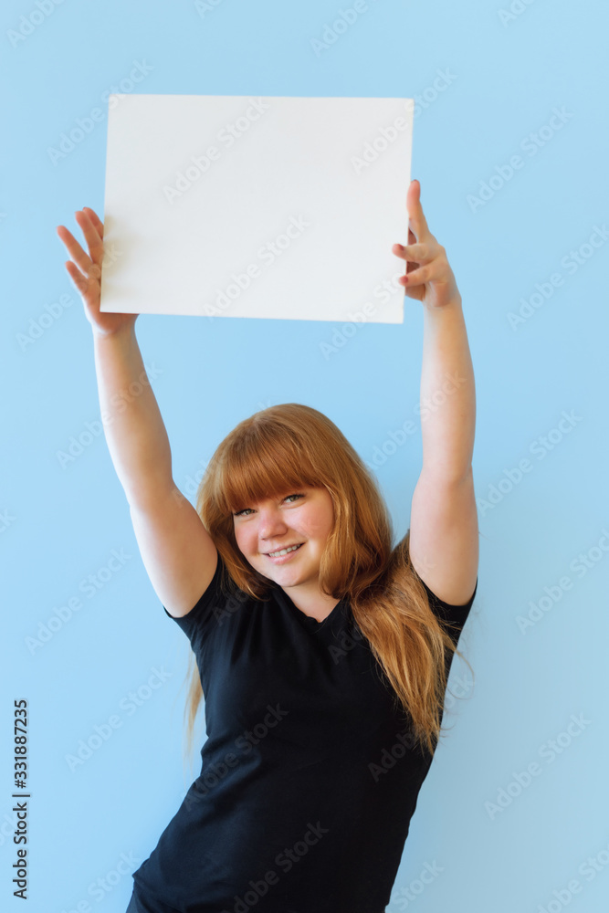 young woman of Caucasian appearance with red hair holds a poster with copyspace with two hands, she smiles, on an isolated blue background, dressed in black casual clothes young woman of Caucasian appearance with red hair holds a poster with copyspace with two hands, she smiles, on an isolated blue background, dressed in black casual clothes