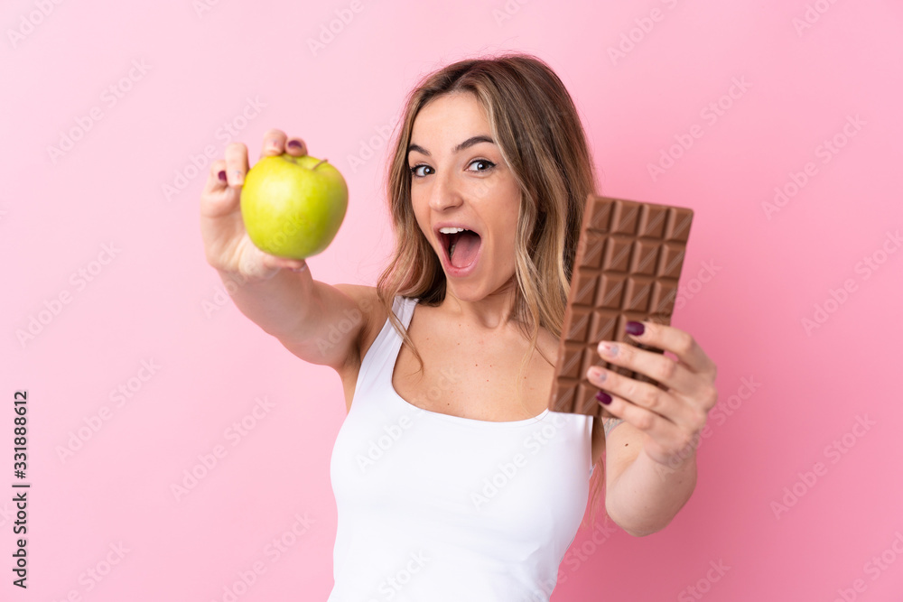 Young woman over isolated pink background taking a chocolate tablet in one hand and an apple in the other Young woman over isolated pink background taking a chocolate tablet in one hand and an apple in the other
