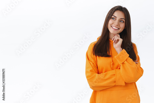 Intrigued and thoughtful caucasian girl in orange hoodie, touch chin tilt head and looking left curious, smiling interested, contemplate cool thing, making choice, standing white background ponder