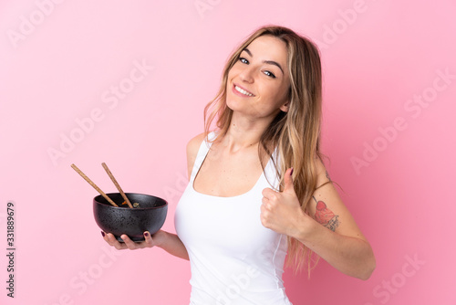 Young woman over isolated pink background with thumbs up because something good has happened while holding a bowl of noodles with chopsticks