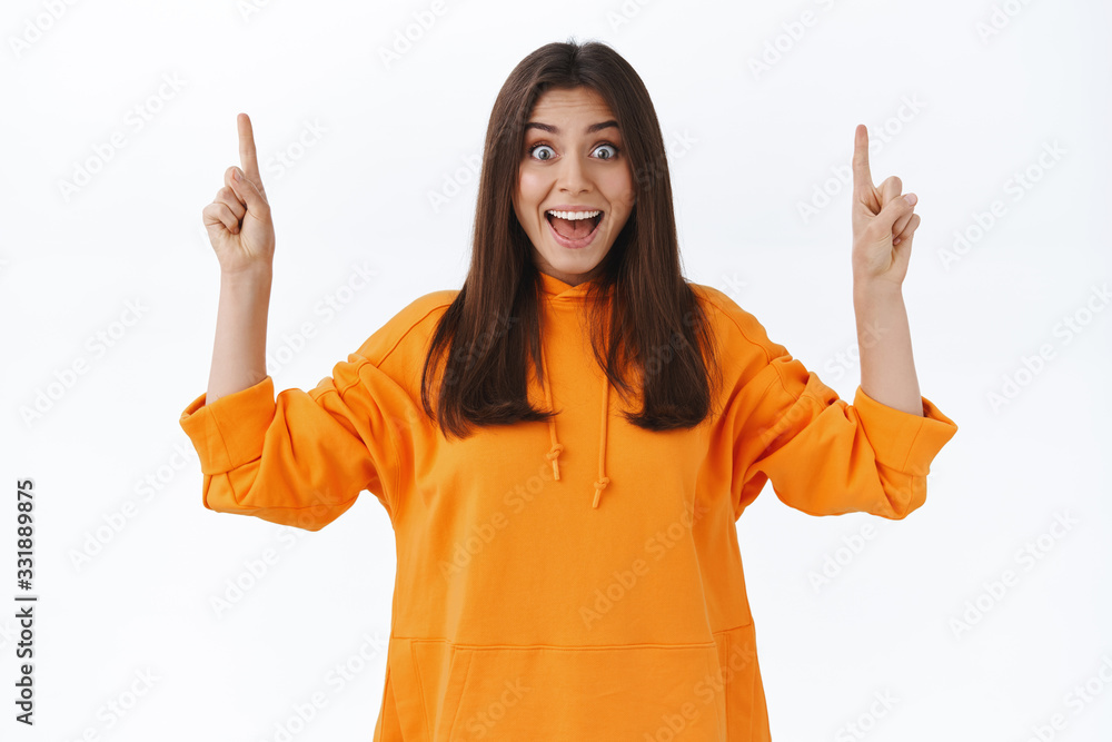Surprised and excited pretty woman in orange hoodie react to cool incredible news, pointing fingers up to tell you about special prices discounts and awesome products in stock, white background