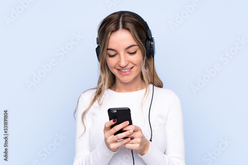 Young woman over isolated blue background listening music and looking to mobile