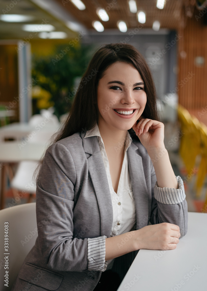 Fototapeta premium Confident businesswoman in modern office, looking at camera, smiling. Portrait of young successful manager sitting at workplace