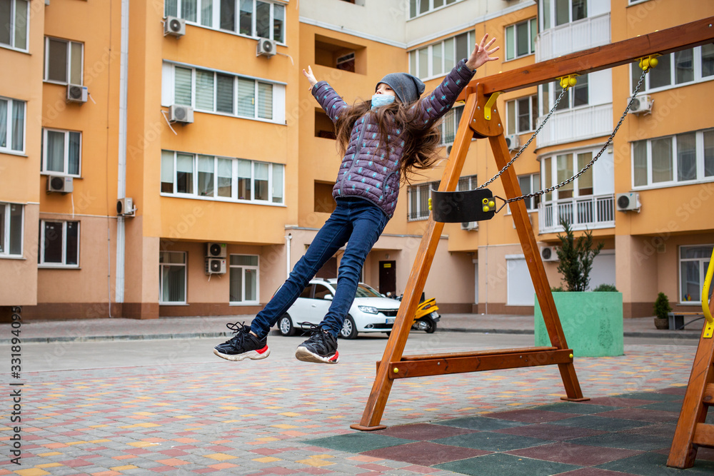 teenage girl in medical mask on her face riding a swing on playground  in background of building in coronavirus quarantine period. European pandemic coronavirus concept