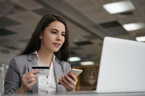 Successful businesswoman holding credit card, using smartphone for online shopping, sitting in office. Freelancer receive payment, check balance. Woman ordering food online, booking tickets