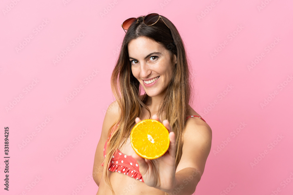 Young blonde woman over isolated pink background in swimsuit and holding an orange Young blonde woman over isolated pink background in swimsuit and holding an orange