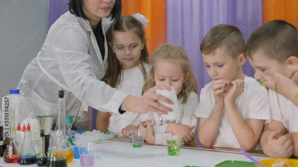 Chemical experiments for children. Woman pours a green substance into a clear glass. Children are surprised. The girl is holding a long green worm. Room is filled with artificial smoke.