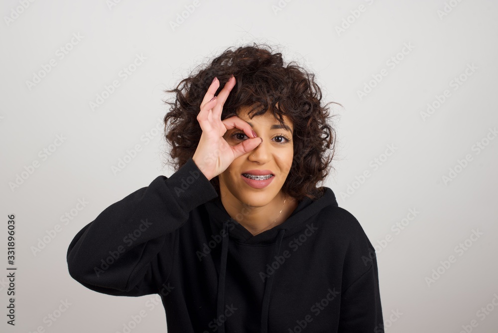Beautiful young blonde woman with happy face smiling doing ok sign with hand on eye looking through finger. Wearing casual clothes and standing. Beautiful young blonde woman with happy face smiling doing ok sign with hand on eye looking through finger. Wearing casual clothes and standing.