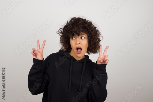 Isolated shot of cheerful woman makes peace or victory sign with both hands, dressed in casual clothes, feels cool has toothy smile, isolated over gray background. People and body language. © Jihan