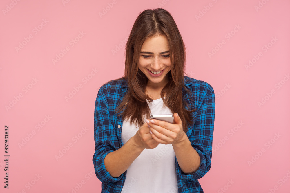 Mobile connection. Portrait of happy brunette girl in checkered shirt reading funny post on social network using cell phone, chatting, typing message. indoor studio shot isolated on pink background Mobile connection. Portrait of happy brunette girl in checkered shirt reading funny post on social network using cell phone, chatting, typing message. indoor studio shot isolated on pink background