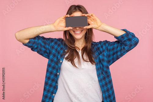 Advertisement on mobile device. Happy girl in checkered shirt covering eyes with smartphone, hiding half face and smiling at camera, expressing joy. indoor studio shot isolated on pink background photo