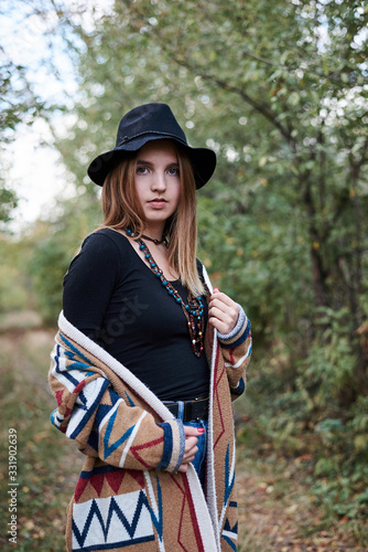 Three-quarter portrait of pretty blond woman in autumn park forest woods. Young girl, wearing black hat, blue mama jeans and colorful cardigan, posing on natural background. Hippie style.
