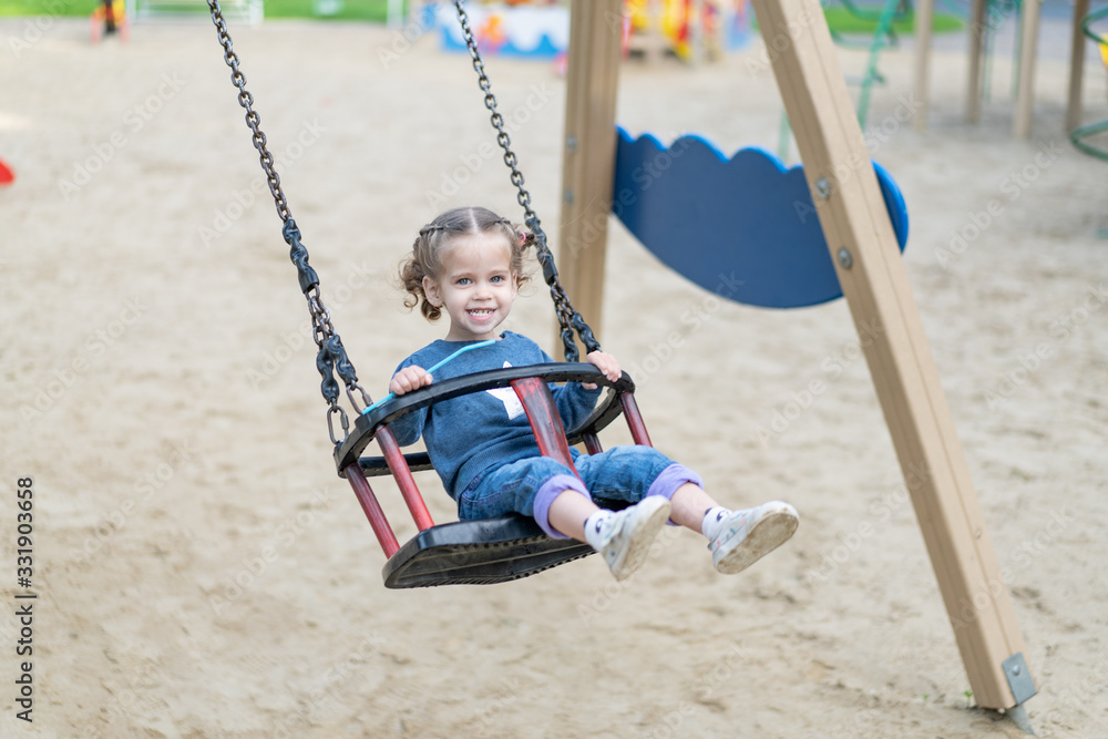 Little Caucasian girl riding swing at playground sunny summer day.