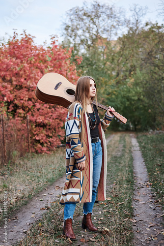 Young blond woman  wearing colorful cardigan and blue jeans  holding acoustic guitar on her shoulder  walking in park woods forest in autumn. Full-length portrait of hippie musician with guitar.